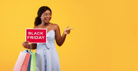 Charming young black woman with shopper bags holding BLACK FRIDAY sign and pointing aside at empty space, orange studio background. African American lady having special offer for sale day