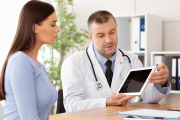 Doctor Appointment At Modern Clinic. Adult specialist holding digital tablet with black empty screen for mockup, showing patient her test results and discussing treatment, sitting at the desk