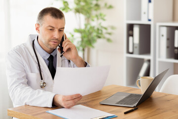 Naklejka premium Medicine Concept. Middle aged male doctor calling his patient, talking on phone and holding medical report