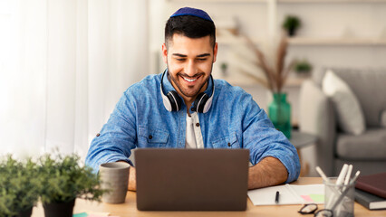 Fototapeta premium Business And Entrepreneurship. Happy Jewish Man In Kippa Working On Laptop Wearing Headphones On Neck Sitting At Workplace In Modern Home Office. Independent Contractor Enjoying Distance Job