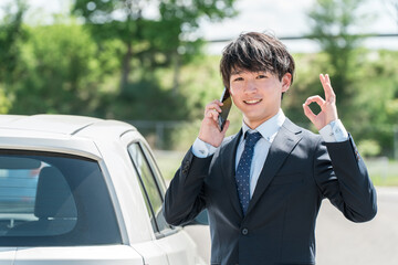 車の前でスマホで通話するスーツ姿の若いアジア人ビジネスマン
