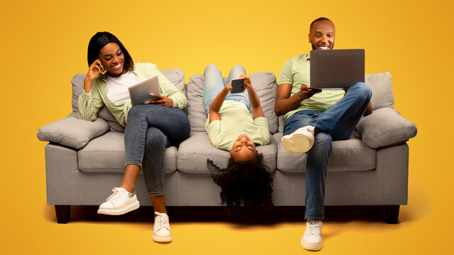 Gadgets addiction. Young black family of three holding and using different electronic devices while sitting on sofa on yellow background. Parents and their daughter with modern gadgets