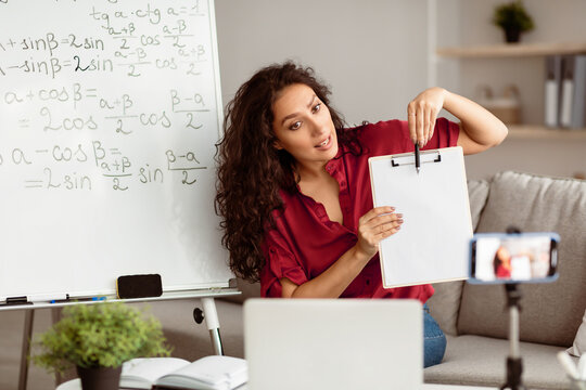 Explaining New Theme. Portrait of young lady tutor having virtual conference, giving online lesson to students using cellphone on tripod. Woman pointing at blank sheet of paper, showing to webcam