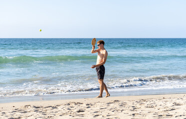 A blonde young man in swimwear and sunglasses enjoying a game of paddle ball on the beach under the sunny sky.