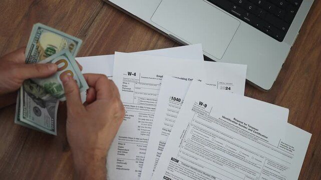 Male accountant spreads out several dollar banknotes in hands counting total cash, person sits at wooden table filled with tax documents and laptop slow motion