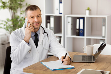 Doctor talking on his smartphone and making notes in a notebook. Medicine professional making phone call with patient and writing in his diary, looking at camera, sitting at desk in his office