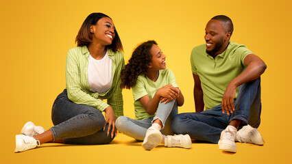 Communication concept. Happy black parents and their daughter talking, sitting on floor of yellow studio background. Positive man having conversation with child and wife