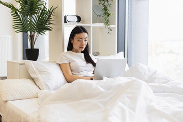 Obraz premium Asian woman in her 20s using laptop in cozy bedroom setting. Relaxed and focused atmosphere with natural daylight and indoor plants adding calmness to home workspace.