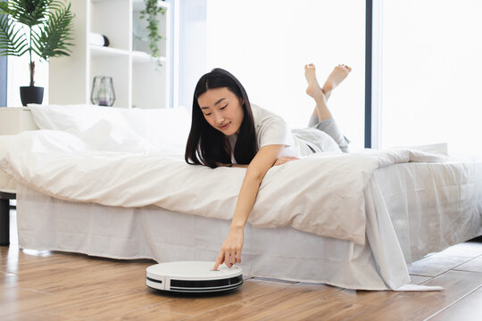Asian woman relaxing on bed while controlling robot vacuum cleaner. Concept of modern technology, convenience, smart home lifestyle. Young adult enjoying clean and tidy living space. - Powered by Adobe