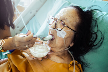Sick Southeast Asian elderly female patient in hospital bed being feed of tender food by her adult daughter. Affection to the sick  family member.