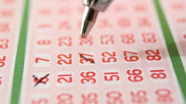 A man marks with a pen numbers 6, 37, 52, 81 on a lottery ticket. The rows of red numbers on the white lottery ticket are arranged vertically. Close-up horizontal video