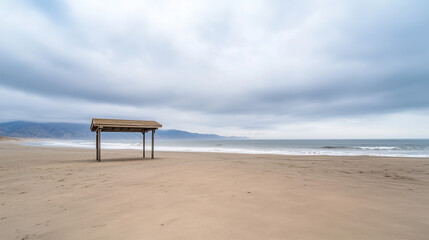 Fototapeta premium Solitary Shelter on a Coastal Beach: A simple wooden beach shelter stands alone on a vast, sandy beach under a brooding, overcast sky. The ocean stretches to the horizon.