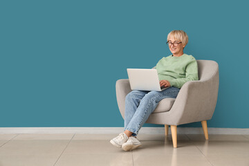 Old woman with laptop sitting in armchair near blue wall