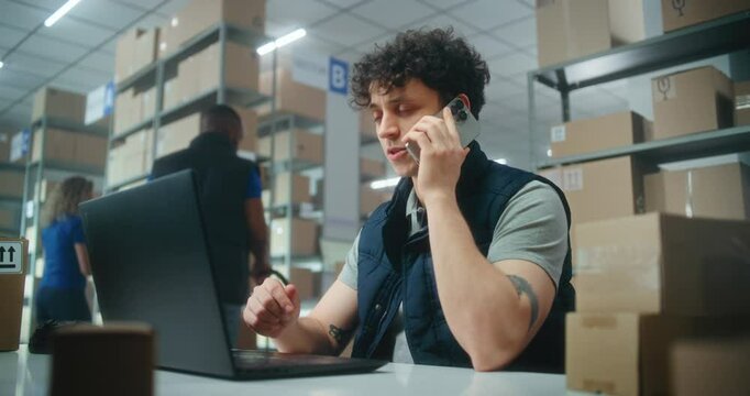 Male logistics coordinator talks to customer on mobile phone, uses laptop computer, works in warehouse of postal service or E-commerce online store. Sorting center employees carrying cardboard boxes.