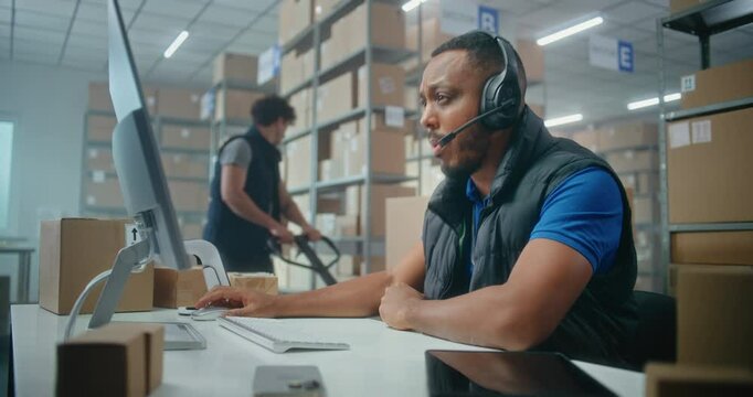 African American logistics coordinator wearing headset chats with customer on call, uses computer, works in post office or E-commerce store storage. Sorting center workers carrying cardboard boxes.
