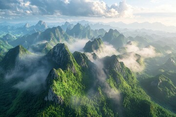 Obraz premium Aerial view of a mountain range is surrounded by clouds and mist during sunrise, towering into the sky. The green peaks stand tall in the sunlight