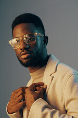 Stylish young man with glasses posing elegantly in a beige coat, showcasing confidence and fashion forwardness against a soft blue background