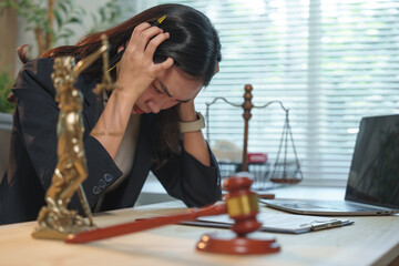 Young asian lawyer woman suffering from a headache and holding her head with hands while sitting at her desk with laptop, gavel, scales of justice and themis statue in law office