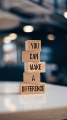 Four wooden blocks stacked vertically on a white surface feature the phrase "YOU CAN MAKE A DIFFERENCE" in bold, dark sans-serif font. A blurred indoor background enhances the focus on the inspiration