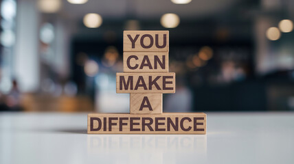 Four wooden blocks stacked vertically on a white surface feature the phrase "YOU CAN MAKE A DIFFERENCE" in bold, dark sans-serif font. A blurred indoor background enhances the focus on the inspiration