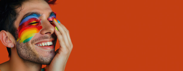 Joyful man with rainbow face paint smiling against vibrant orange background, space for text or design. Embracing colors and pride with a cheerful, expressive gesture that radiates happiness.