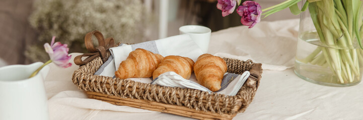 Fresh croissants in a wicker basket on the table next to a vase of lilac tulips.