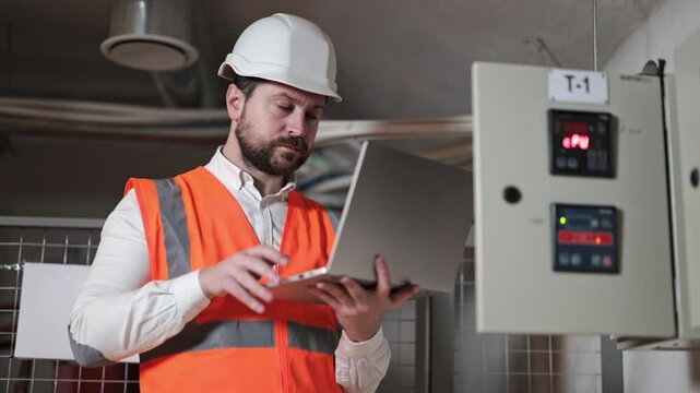 Serious electrical engineer with a beard and an orange vest types on a laptop while examining electrical control panel. Focused industrial worker using a laptop in front of an electrical switchboard.