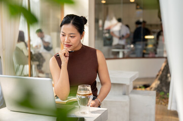 A beautiful, focused Asian woman reads something on her laptop screen, working remotely from a cafe.