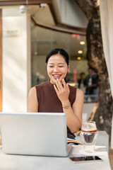 A happy, confident Asian woman is working remotely from a cafe, participating in an online meeting.