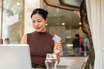 A satisfied Asian woman holds her credit card while shopping online on her laptop, sitting in a cafe