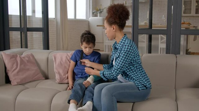 Caring African American mother adjusting arm sling bandage to child injured upper arm to heal fracture received by preadolescent black son, while family sitting on sofa in domestic room.