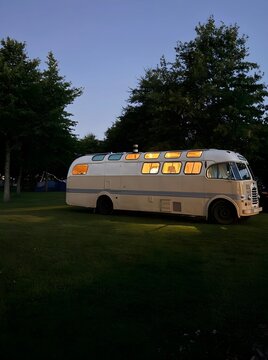 Vintage camper bus glowing warmly in the twilight, surrounded by trees on a grassy campsite, evoking nostalgia and a serene outdoor ambiance.