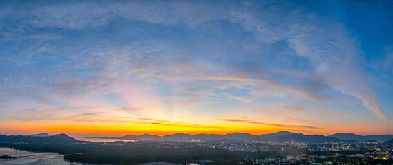 A stunning panoramic view of a coastal city at sunset at Phuket city, showcasing vibrant hues of orange and blue in the sky, with mountains, and urban lights creating a harmonious blend of natural.
