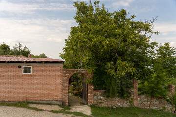 A brick house with a small archway leading to a courtyard. The courtyard is surrounded by a brick wall and has a tree in it. The sky is cloudy and the house is located on a hill