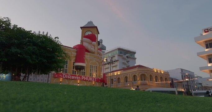 Phuket,Thailand-December,25,2024: A vibrant scene featuring a historic yellow clock tower adorned with giant Santa Claus and Mrs. Claus decorations during a dramatic evening sky. Happy New Year 2025