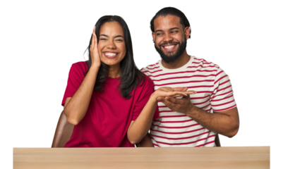 Seated young Latino couple at table holds copy space on a palm, keep hand over cheek. Amazed and delighted.