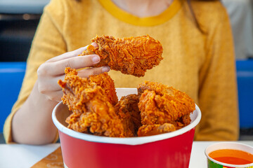 Girl holding a fried chicken drum stick with a bucket full of chicken pieces in a cafe restaurant lifestyle environment.