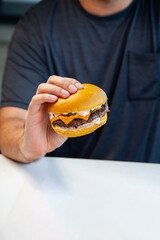 Man wearing black T-shirt holding seven ounces of prime beef burger with cheddar cheese dill pickles and delicious sauce inside a cafe restaurant.