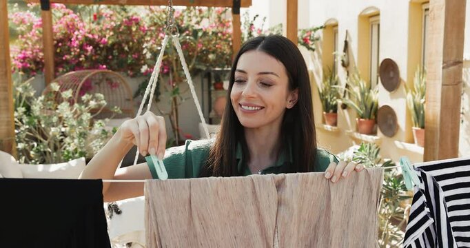 Woman hangs clothes on clothesline, drying garment in fresh air, standing outdoors in backyard garden filled with vibrant plants and flowers during sunny summer day, smile, do routine household chores