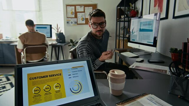 POV of man making marketing presentation on laptop, sitting at desk, while discussing questions with his male colleague in modern open space office