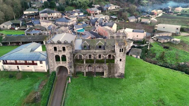 Aerial shot of Arnedo Castle, a historic stone fortress with arches and towers, surrounded by green fields and a small village in Valdeorras.