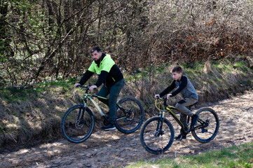 Obraz premium A man and boy cycle along a dirt path amidst trees. Sunlight filters through the branches, illuminating the scene. They appear to be enjoying a leisurely ride together.
