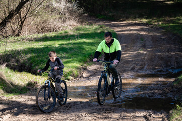 A man and a boy on bicycles navigate a muddy path in a wooded area. They are crossing a small,...