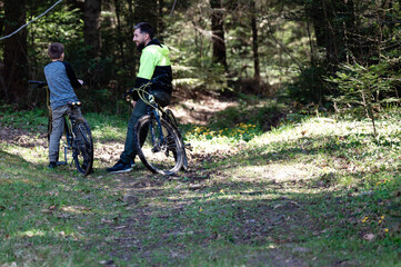 A man and a boy take a break from their bike ride. They're sitting on their bikes, talking, amidst the tranquil forest setting. Sunlight filters through the trees.
