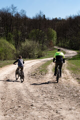 Two figures on bicycles ride along a winding dirt path. Trees and greenery line the road, creating a picturesque setting. A sunny day enhances the outdoor activity.