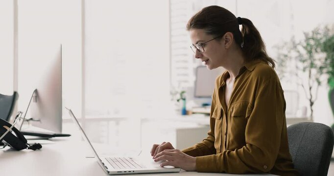 Young 30s attractive woman wear glasses working on laptop seated at desk in contemporary office, responds on e-mails, analyzing data, drafting documents, or conducting research. Business, technology