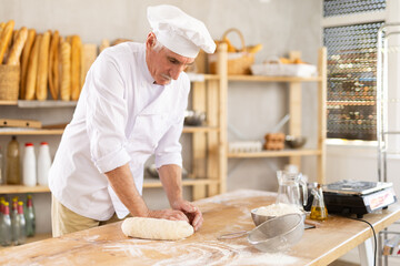 Elderly male baker preparing raw yeast dough on kitchen table in bakery