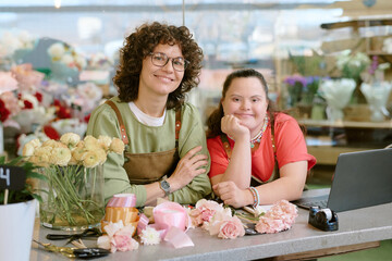 Medium close up portrait of two adorable florists with brown velvet aprons leaning on table with devices, satin ribbons and aromatic flowers