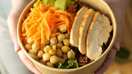 Woman holding nutritious meal poki bowl featuring grilled chicken breast, chickpeas, shredded carrots, avocado slices, brown rice
