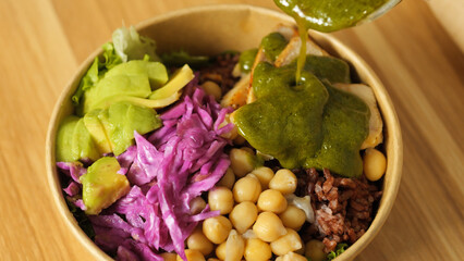 Chef pouring vibrant pesto sauce over a colorful buddha bowl filled with grilled chicken, avocado, chickpeas, red cabbage, and brown rice for a nutritious meal